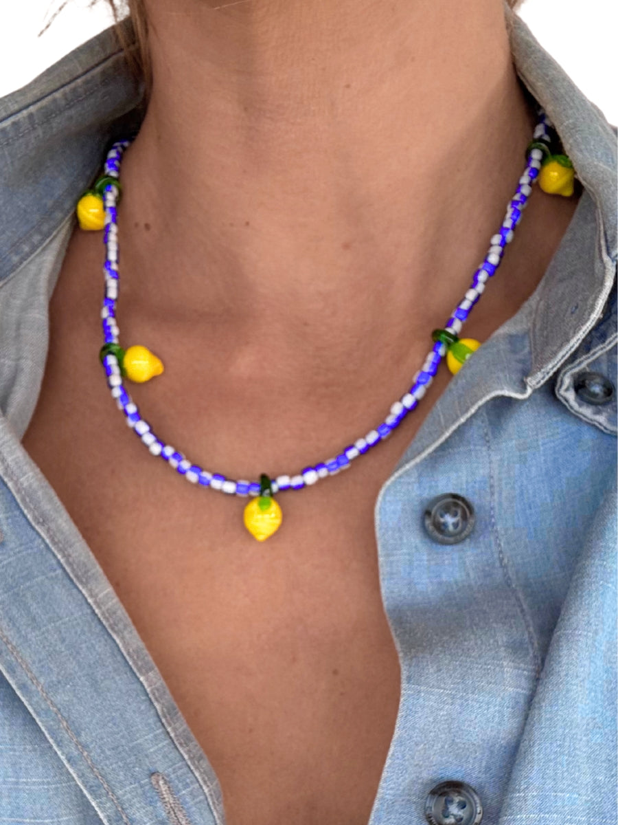A close-up of a person wearing a beaded necklace with yellow lemon-shaped beads and blue beads, styled with a light blue denim shirt.