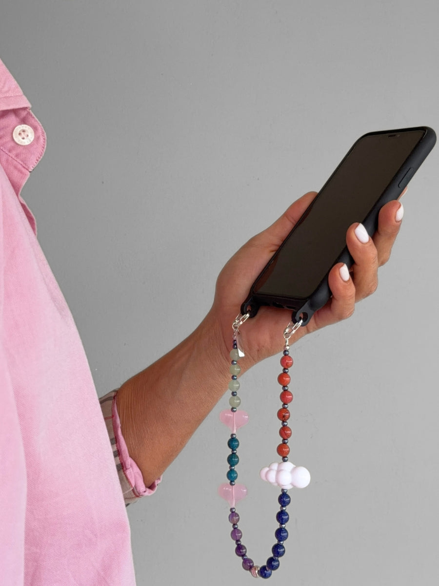A close-up of a woman's hand holding a black smartphone with a colorful beaded phone strap. The strap features multi-colored round beads with small pink hearts and white cloud charms.