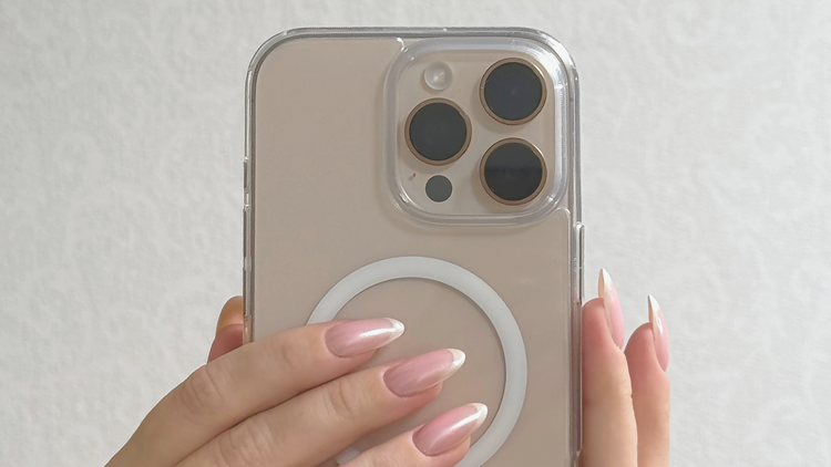 Close-up of a hand with elegant pink nails holding a clear MagSafe phone case on a beige smartphone, showcasing a stylish and modern phone accessory.