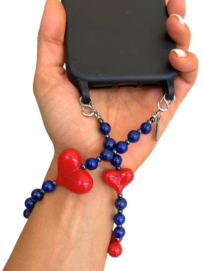A hand holding a black smartphone with a blue and red beaded wrist strap featuring two red heart charms and silver metal clips, shown against a plain white background.