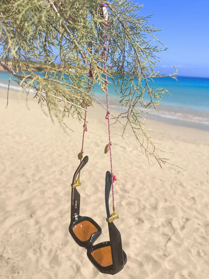 Black sunglasses hang from a tree with a colorful boho neck strap, set against a beach with the ocean in the background.
