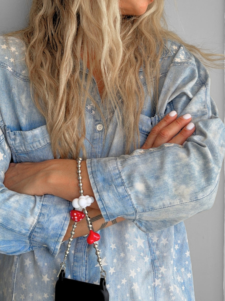 A close-up of a person wearing a light blue denim shirt with a white star pattern, holding a black shoulder bag with a beaded strap featuring red and white cloud-shaped charms.