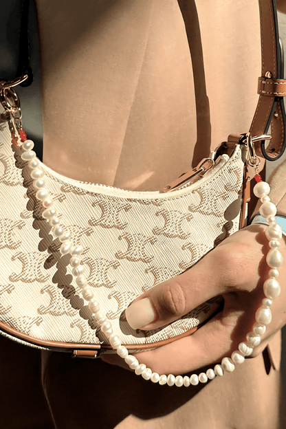 Hand holding a beige handbag with a pattern, wearing a pearl bag charm