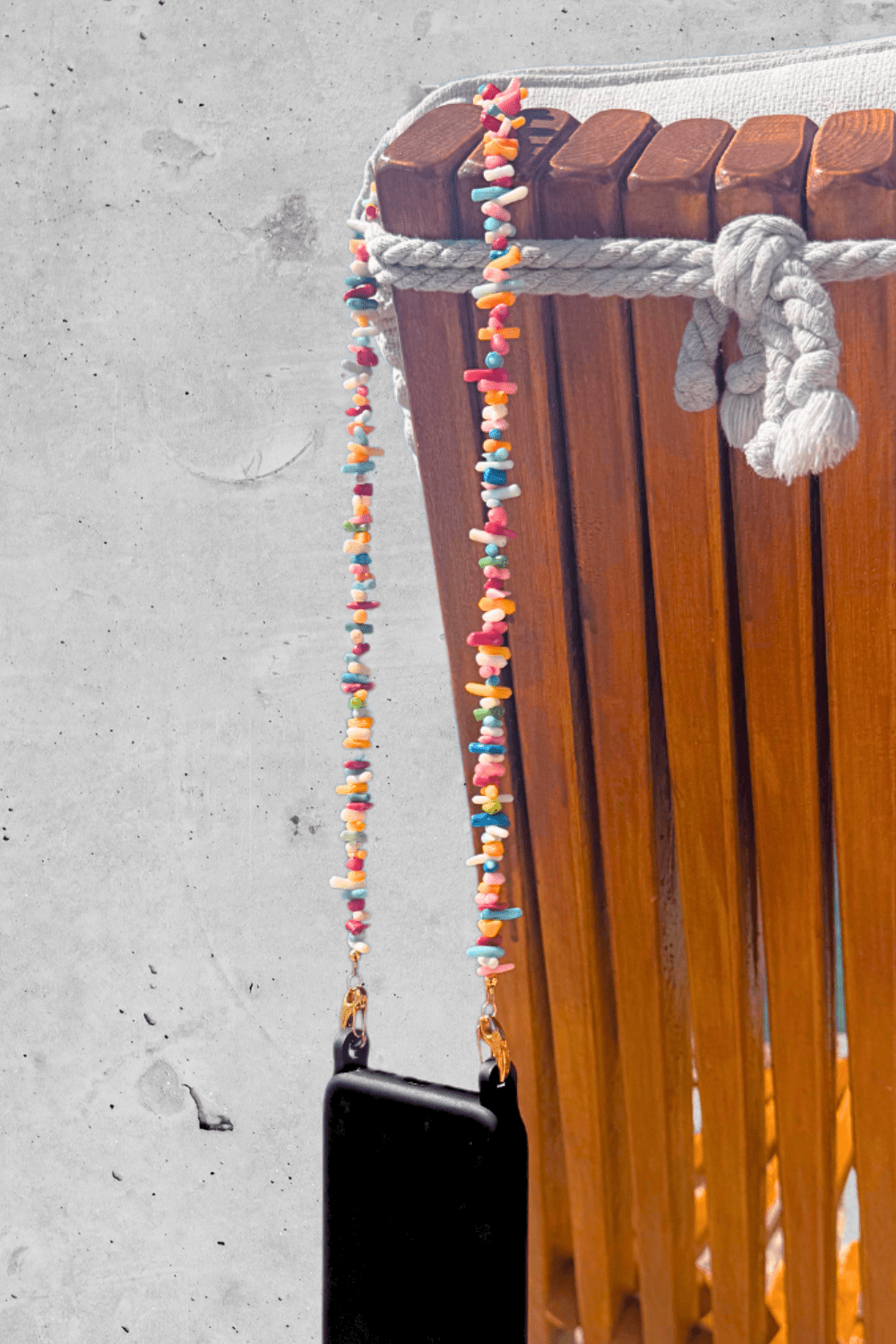 Colorful beaded phone and bag strap hanging on a wooden chair against a concrete wall.
