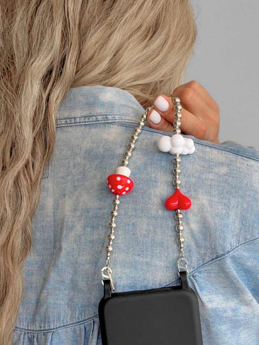 A close-up of a person wearing a light blue denim shirt, holding a silver beaded phone strap with a red and white mushroom charm and a red heart charm. A black phone case is attached to the strap.