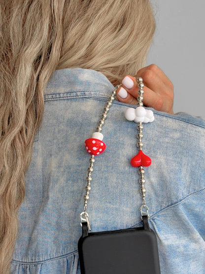 A close-up of a person wearing a light blue denim shirt, holding a silver beaded phone strap with a red and white mushroom charm and a red heart charm. A black phone case is attached to the strap.