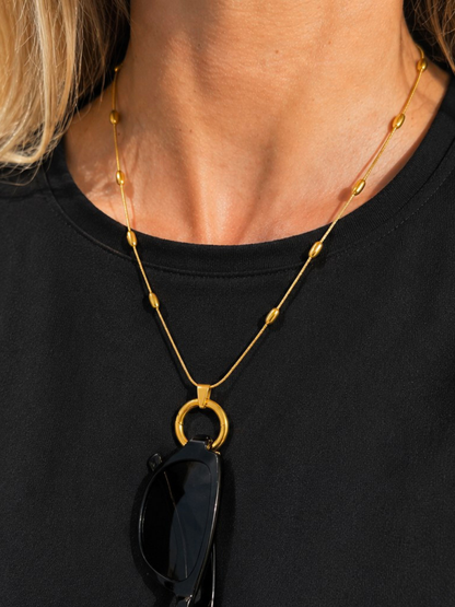 A close-up of a model wearing a gold necklace with a large circular ring pendant and small bead accents, styled over a black t-shirt with sunglasses hanging from the pendant.
