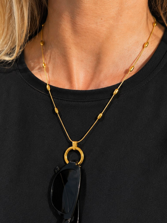 A close-up of a model wearing a gold necklace with a large circular ring pendant and small bead accents, styled over a black t-shirt with sunglasses hanging from the pendant.