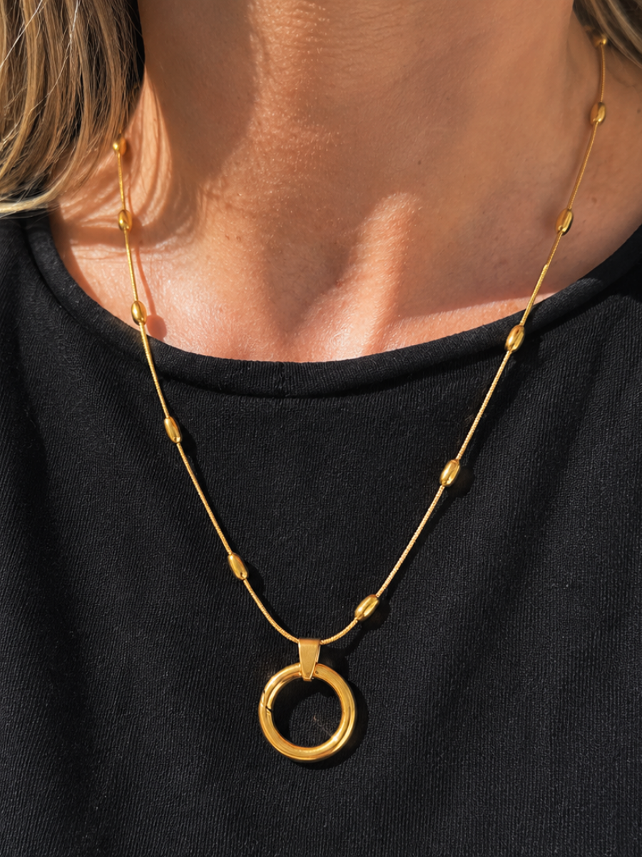 A close-up of a person wearing a delicate gold necklace with small bead accents and a large circular ring pendant, paired with a black top.