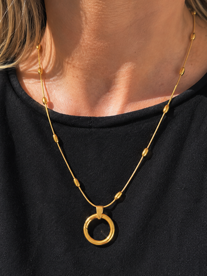 A close-up of a person wearing a delicate gold necklace with small bead accents and a large circular ring pendant, paired with a black top.