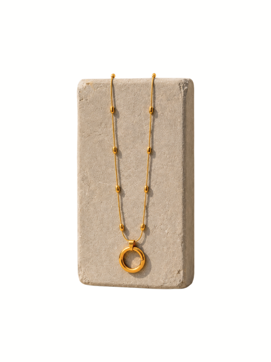 A gold necklace with a textured circular pendant and small bead accents on a chain, displayed on a rectangular concrete block against a white background.