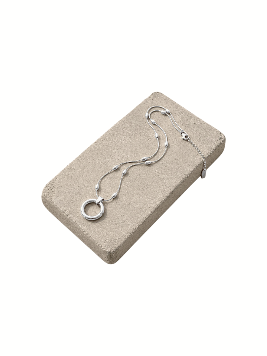 A delicate silver-toned chain necklace with a circular ring pendant and small bead accents, displayed on a rectangular beige jewelry presentation block against a white background.