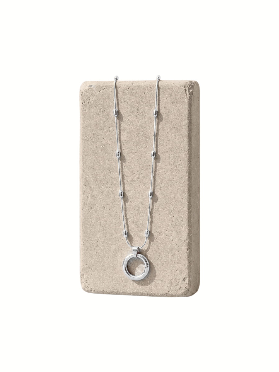 A silver beaded necklace with a circular pendant, displayed on a rectangular beige stone block against a white background.