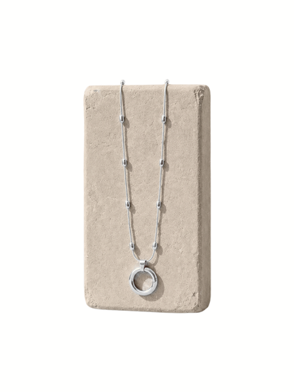 A silver beaded necklace with a circular pendant, displayed on a rectangular beige stone block against a white background.