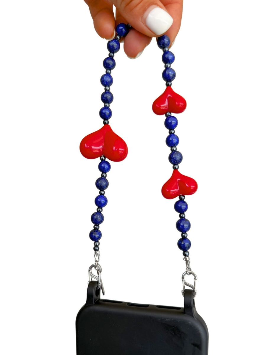 A phone strap with blue beads and three red heart charms, held by a hand with white nail polish against a white background.