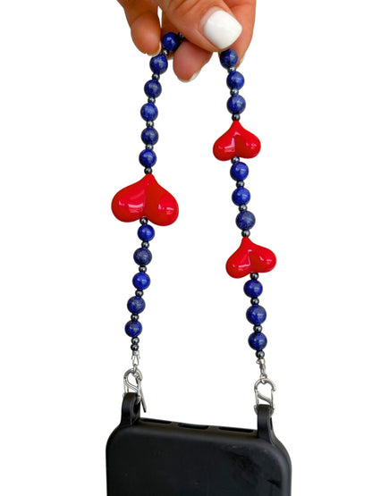 A phone strap with blue beads and three red heart charms, held by a hand with white nail polish against a white background.