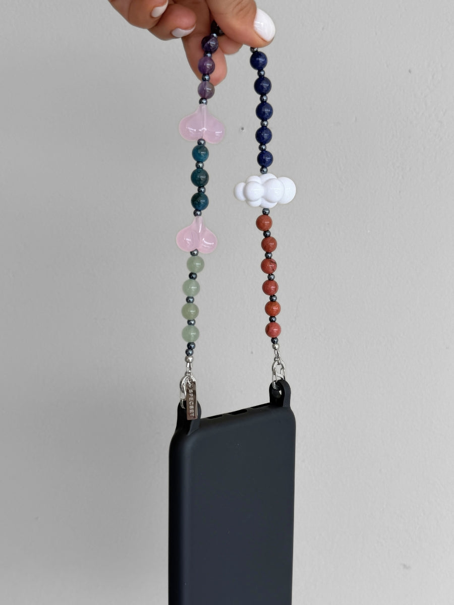 A dark gray phone case with a decorative beaded wrist strap is held by a hand. The strap features multicolor gemstone beads, a white cloud charm, and pink hearts.