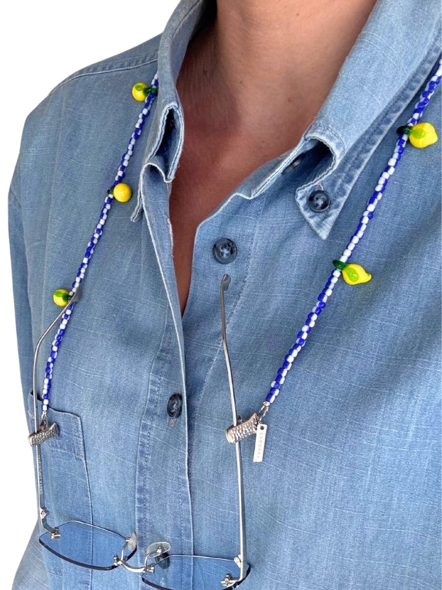 A close-up of a person wearing a light blue denim shirt, with a blue and white beaded necklace featuring small yellow lemon-shaped beads and silver metal clips holding sunglasses.