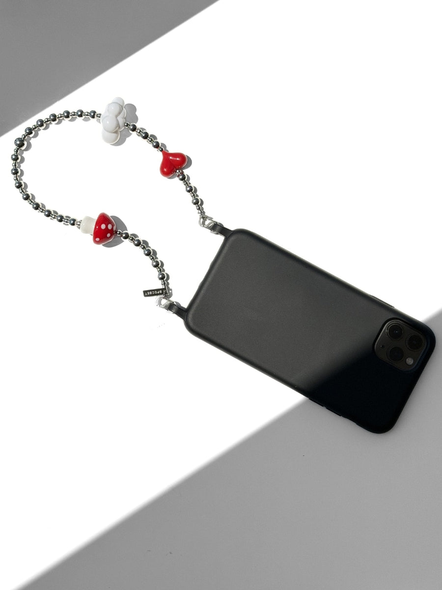 A black smartphone case with an attached silver chain strap featuring red heart-shaped beads, a white flower charm, and a small white mushroom charm, displayed on a white surface with a gray shadow.