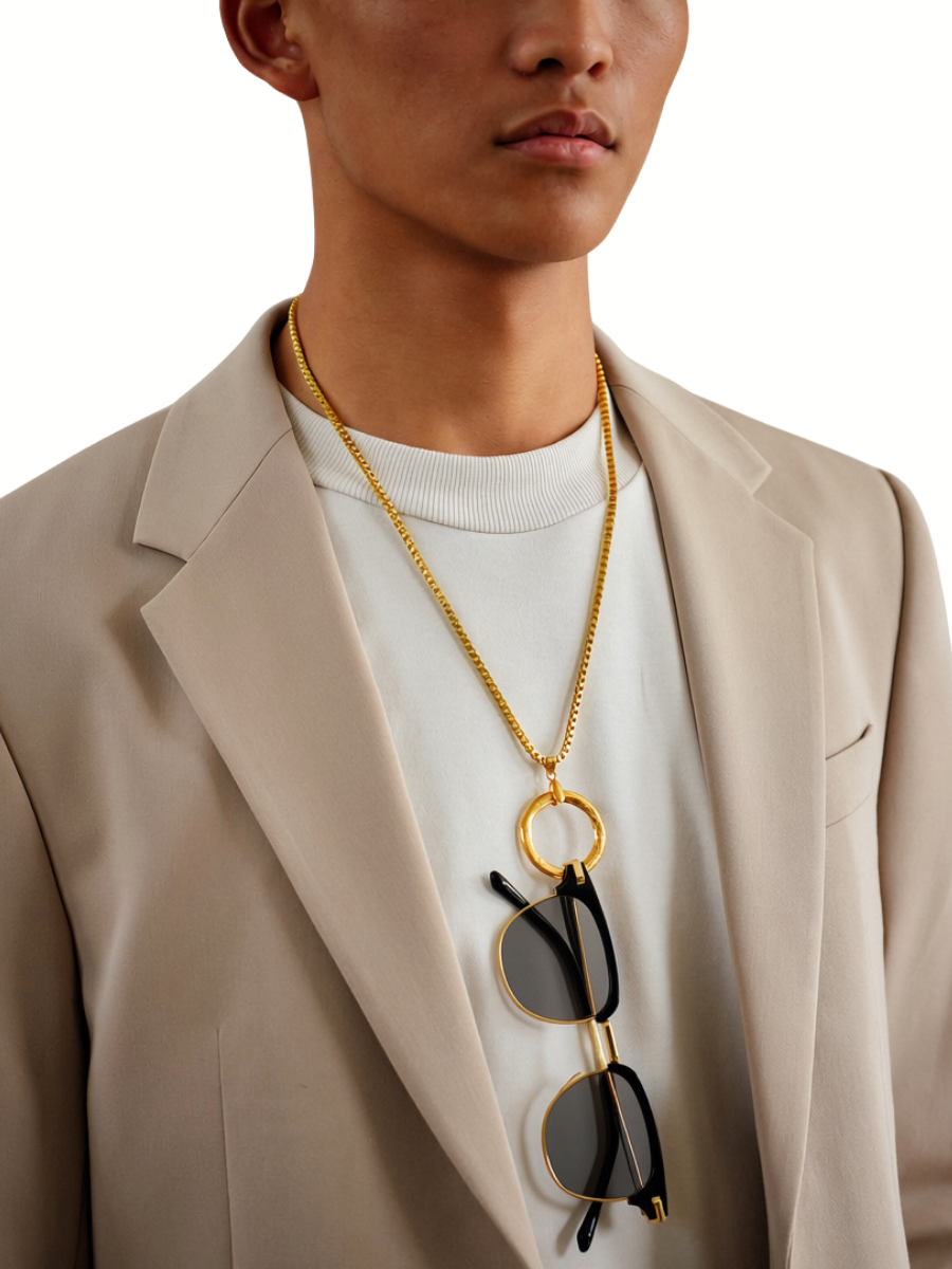 A close-up of a model wearing a gold chain necklace with a large circular ring pendant and sunglasses hanging from it, styled over a white t-shirt and beige blazer.