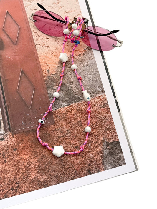 A pink beaded sunglasses strap with white ceramic beads, shown attached to pink sunglasses on a printed card against a white background.