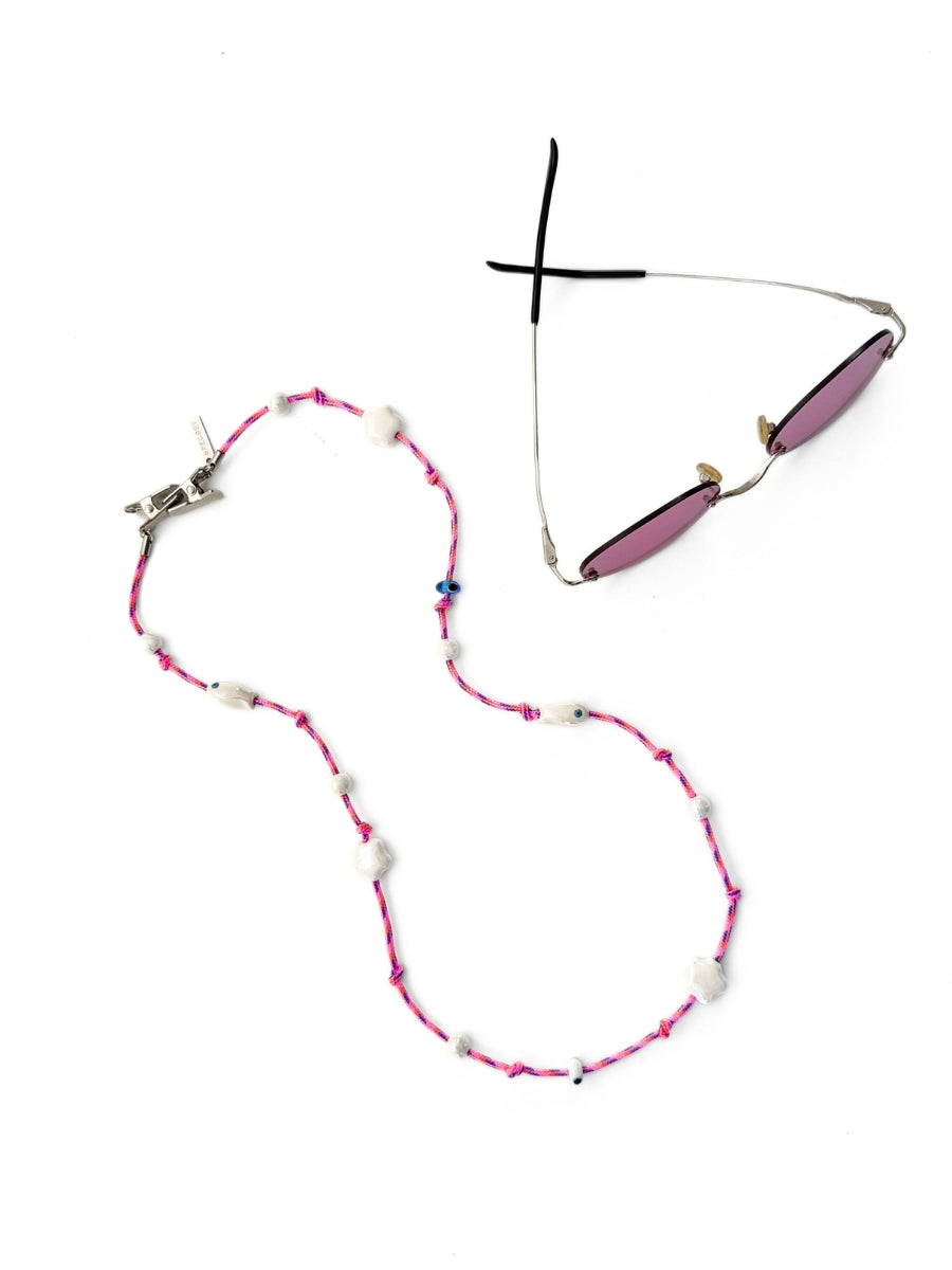 A pair of sunglasses with pink lenses and a silver metal frame, displayed next to a pink and white beaded necklace on a plain white background.