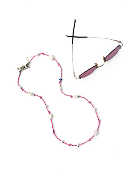 A pair of sunglasses with pink lenses and a silver metal frame, displayed next to a pink and white beaded necklace on a plain white background.