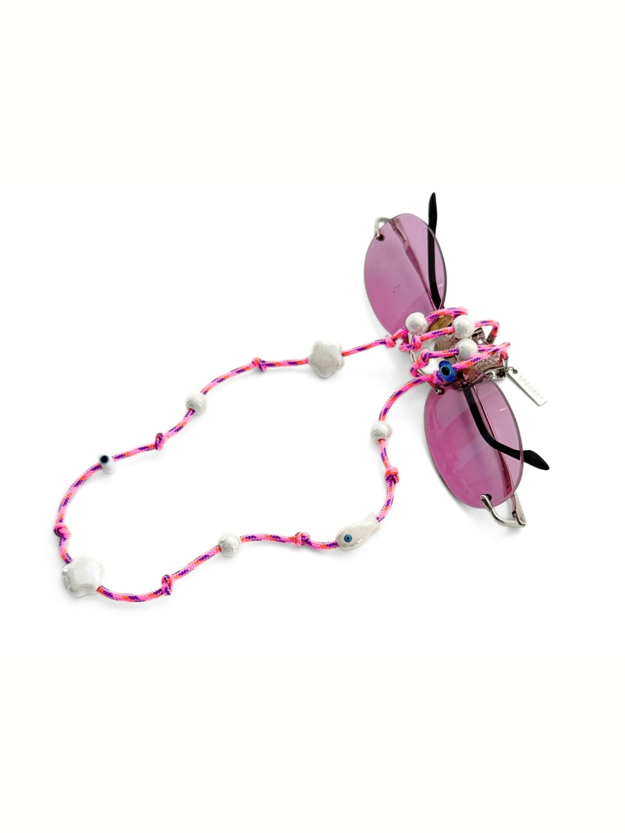 Pink braided eyewear strap with white ceramic beads and evil eye charms, wrapped on a pair of pink sunglasses against white background.
