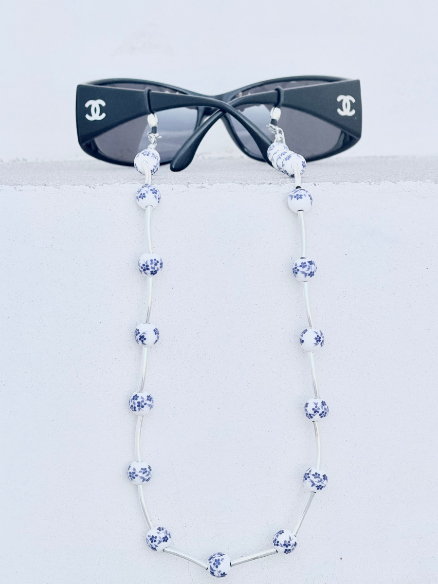 A pair of black sunglasses with a long silver strap made of blue-and-white patterned beads, displayed against a light gray background.