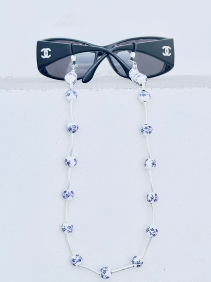 A pair of black sunglasses with a long silver strap made of blue-and-white patterned beads, displayed against a light gray background.