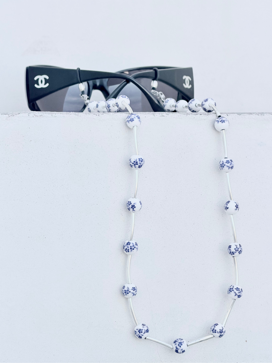 A beaded necklace with white beads featuring blue floral patterns, draped over a white surface with black sunglasses resting on top. The sunglasses have visible Chanel logos on the temples.