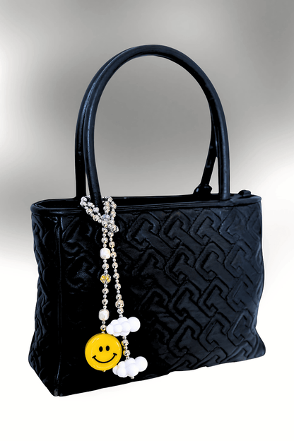 SMILEY bag charm clipped to the handle of a black tote bag, showing its cheerful smiley face and cloud details