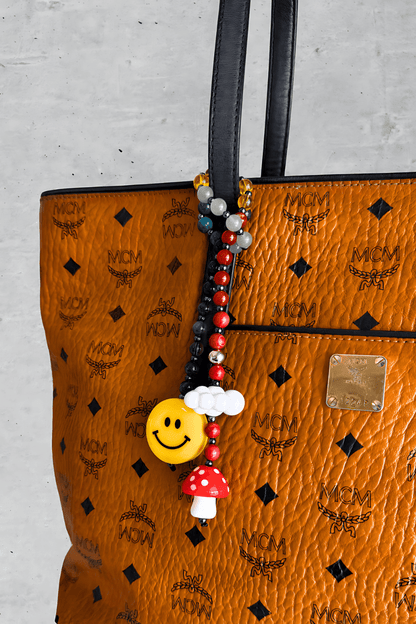 Colorful beaded bag charm with smiley face, mushroom, and cloud - attached to luxury brown MCM handbag handle.

