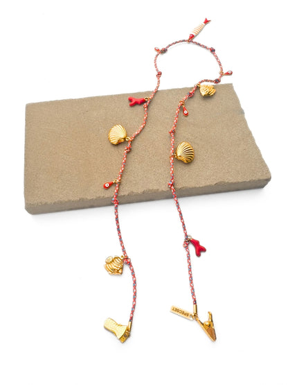 A beach-inspired eyewear strap with gold seashell and evil eye charms, draped over a rectangular stone block on a white background.