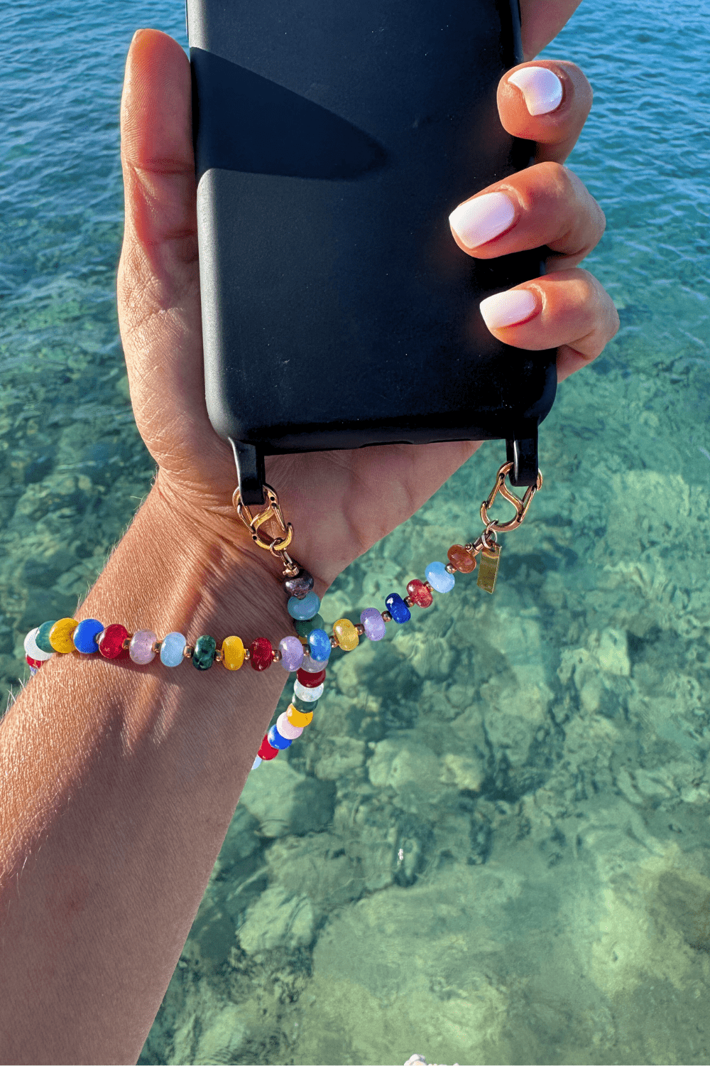 Woman holding a phone over clear blue water, wearing the Vivid Way chain securely as a wrist phone strap, showcasing the vibrant colors.
