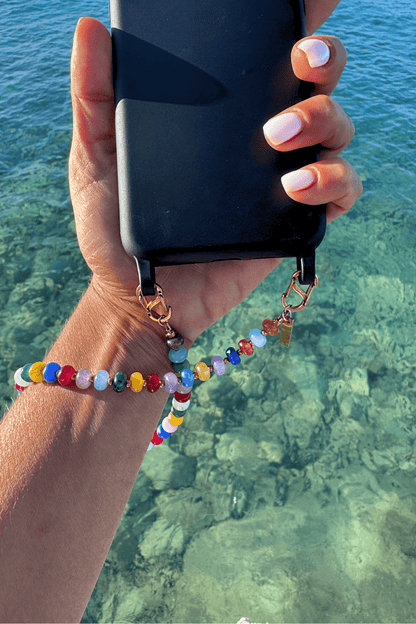 Woman holding a phone over clear blue water, wearing the Vivid Way chain securely as a wrist phone strap, showcasing the vibrant colors.