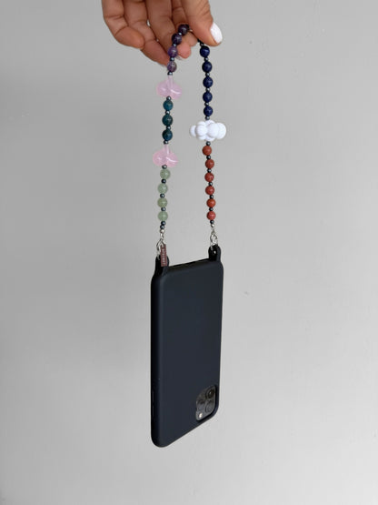 A hand holding a black phone case with a decorative beaded wrist strap featuring pink hearts and white cloud charms against a neutral gray background.