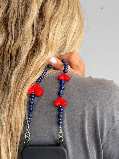 A close-up lifestyle shot of a woman with long blonde hair wearing a gray shirt, holding a phone strap with dark blue beads and red heart charms over her shoulder.