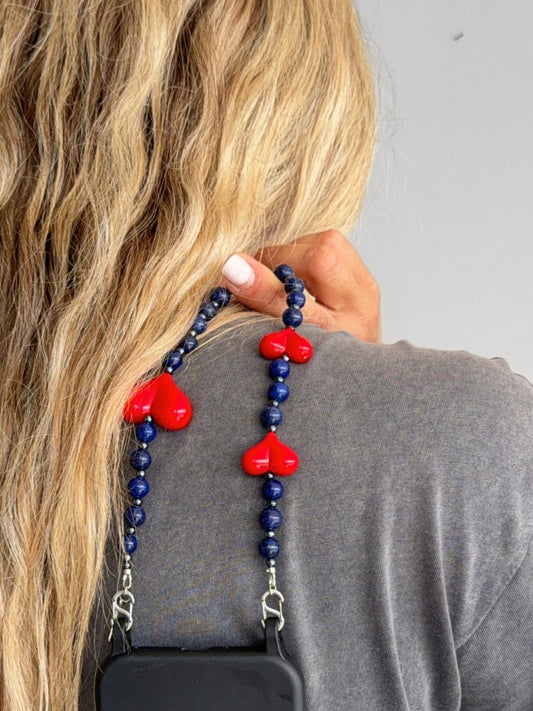 A close-up lifestyle shot of a woman with long blonde hair wearing a gray shirt, holding a phone strap with dark blue beads and red heart charms over her shoulder.