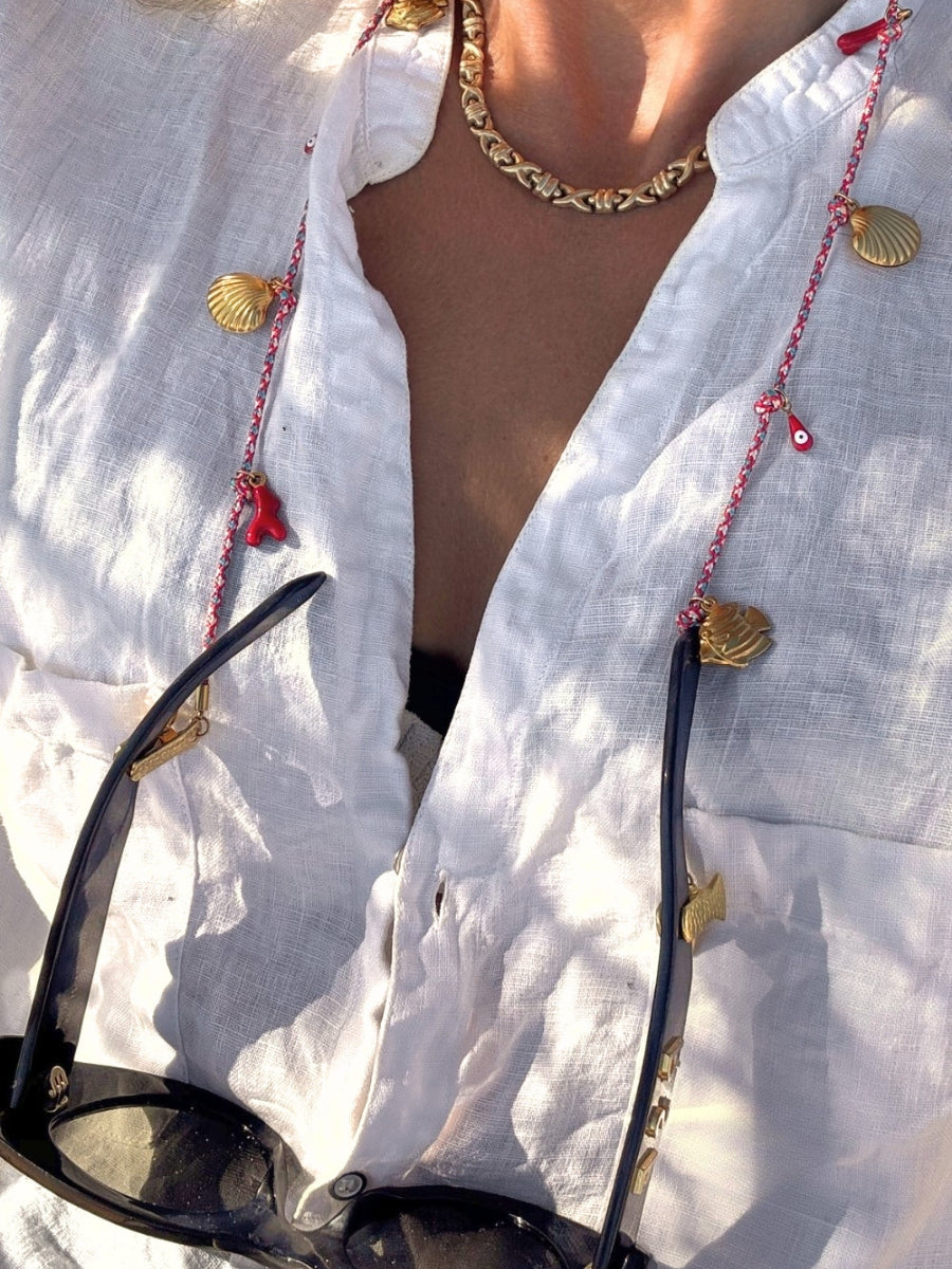 A woman with a white shirt wearing a red neck strap decorated with gold shell charms and small evil eyes. Black sunglasses are draped over the chest.